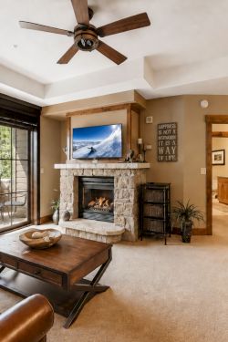 A cozy living room with a stone fireplace, flat-screen TV, ceiling fan, and large glass doors to a patio view.