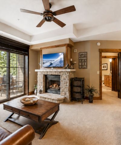 A cozy living room with a stone fireplace, flat-screen TV, ceiling fan, and large glass doors to a patio view.