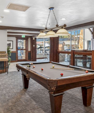 An indoor billiards room with a full-size pool table, cue balls and racked balls, plus lamps, chairs, and glass barriers.