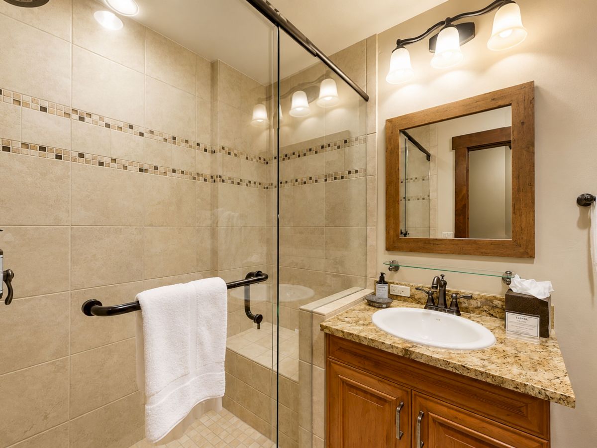 A bright bathroom with a glass shower, tiled walls, a granite vanity and sink, and wall-mounted lights above a wooden mirror.