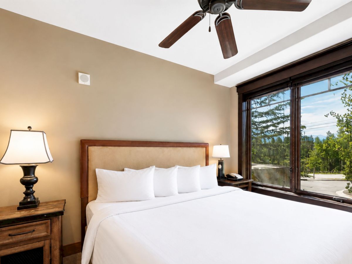 A bright hotel bedroom with a ceiling fan, lamp, neatly made bed, and large window showing greenery and distant mountains.