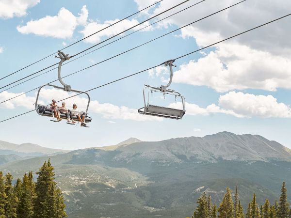 A mountain chairlift carries passengers across scenic peaks under a blue sky and clouds.