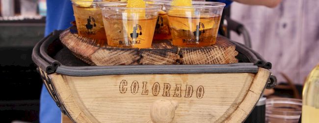 Four clear cups of orange cocktail/punch with lemon slices sit on a wooden barrel at a distillery tasting event.