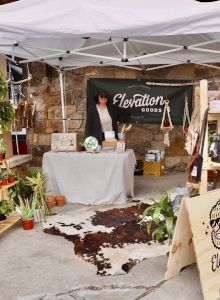 An outdoor vendor booth for Elevation Goods at a market, with plants, bags, and a vendor table under a white canopy.