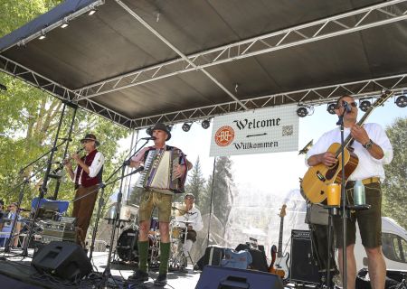 Outdoor festival stage where musicians play accordion and guitar under lights and a &ldquo;Willkommen&rdquo; banner.