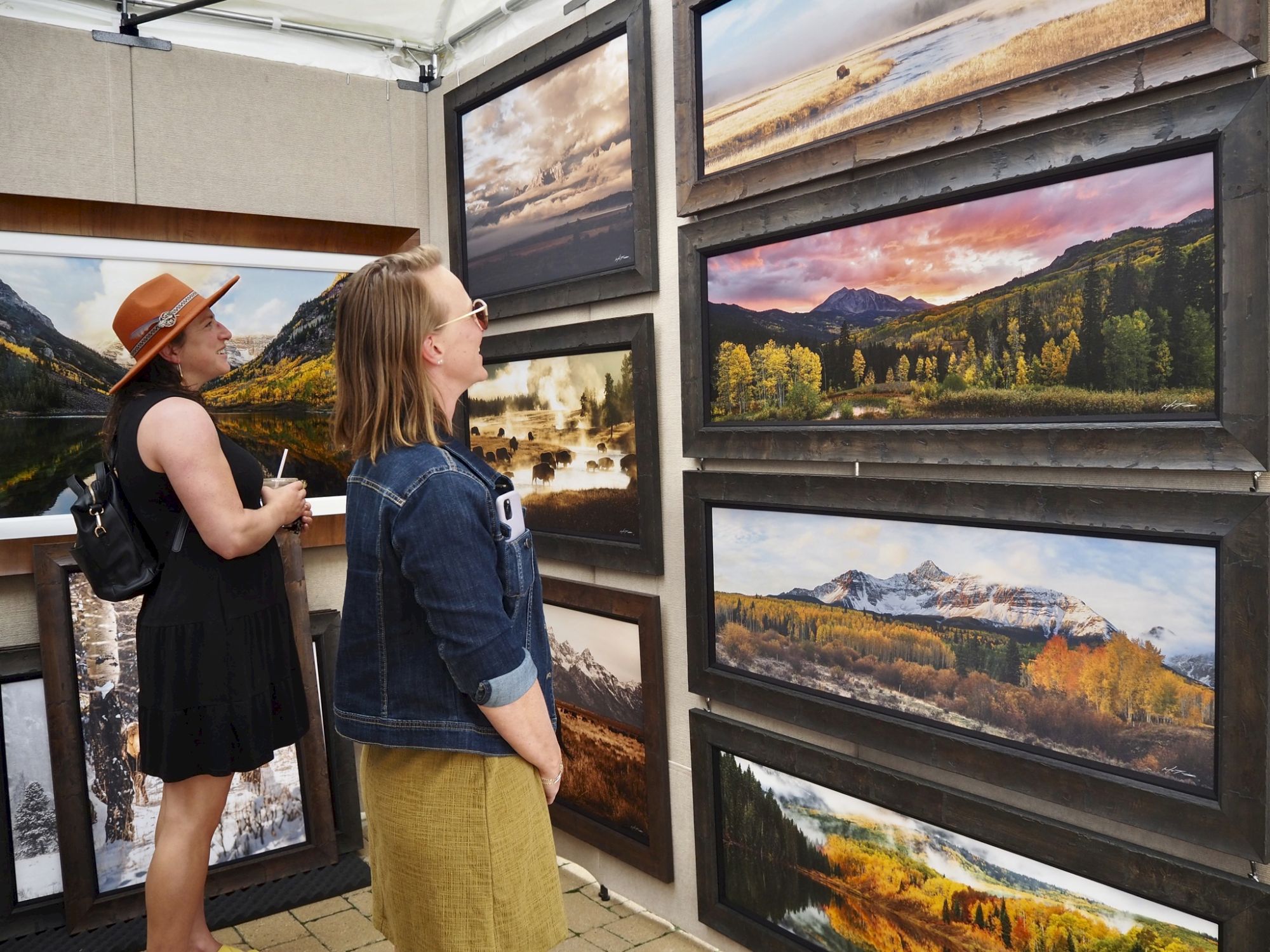 Two women browse framed mountain landscape photos in a gallery, admiring vivid scenery.