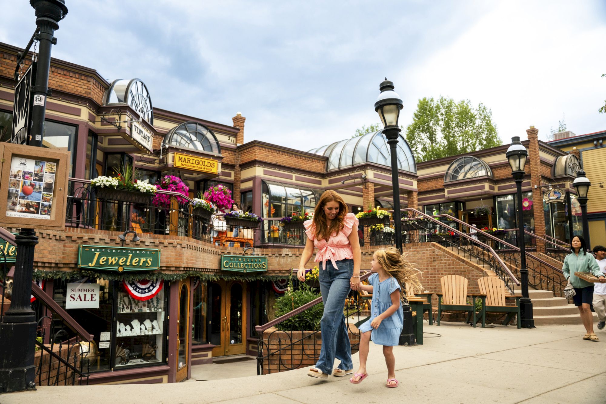 Outdoor shopping arcade with &ldquo;Jewelers&rdquo; storefronts, flowers and glass roofs, as a woman walks with a little girl.